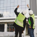 Multi racial builders standing outdoors, back view. Wearing uniform, talking about new glass building. Working on the poject. City infrastructure. Black man.